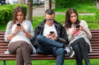 Three young men and women sitting on park bench looking at their phones